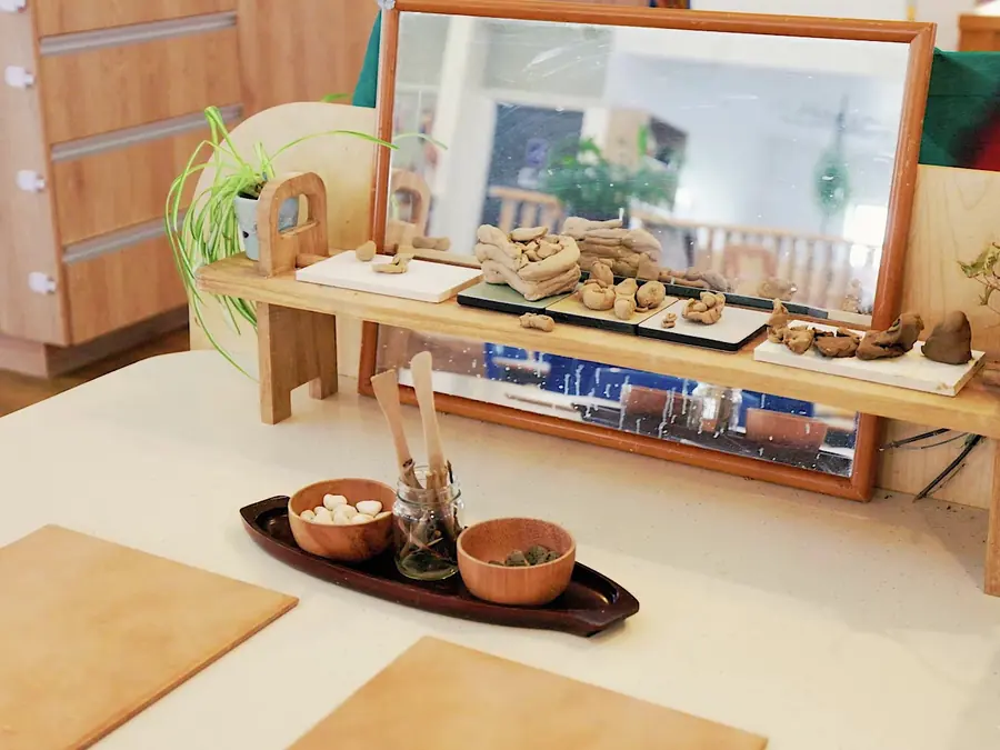 Clay pieces on a low shelf in front of a mirror, with wooden tools and bowls of stones on the table below.