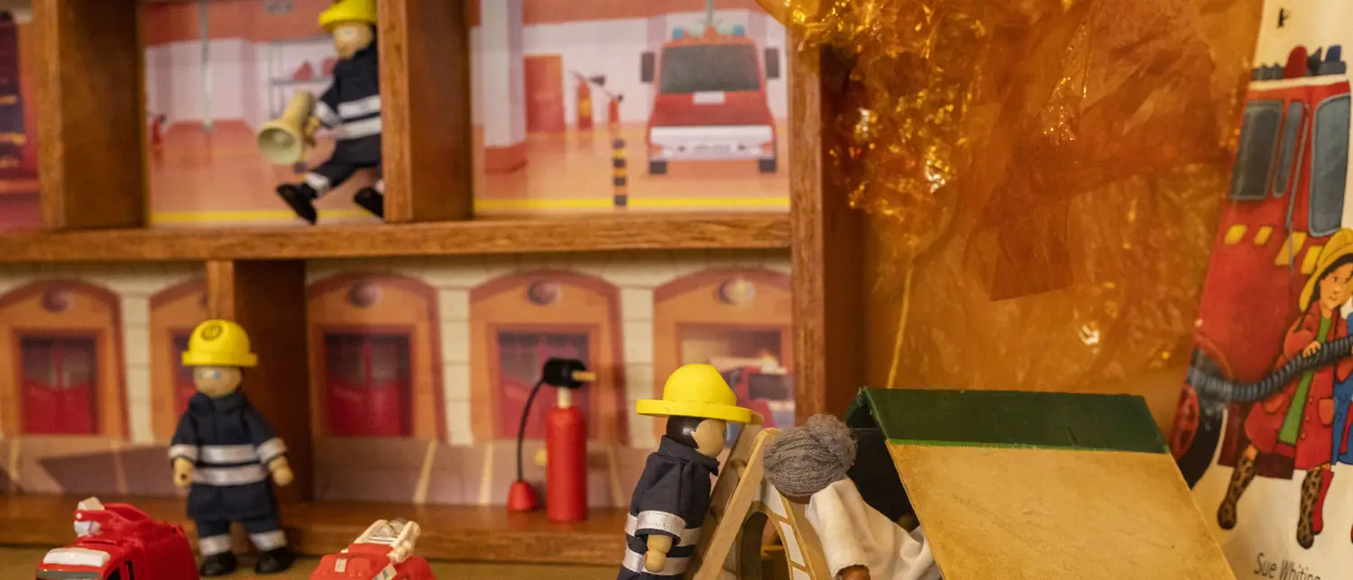 A small-world fire station on a table: wooden firefighters in yellow helmets, red toy fire engines and orange cellophane flames.