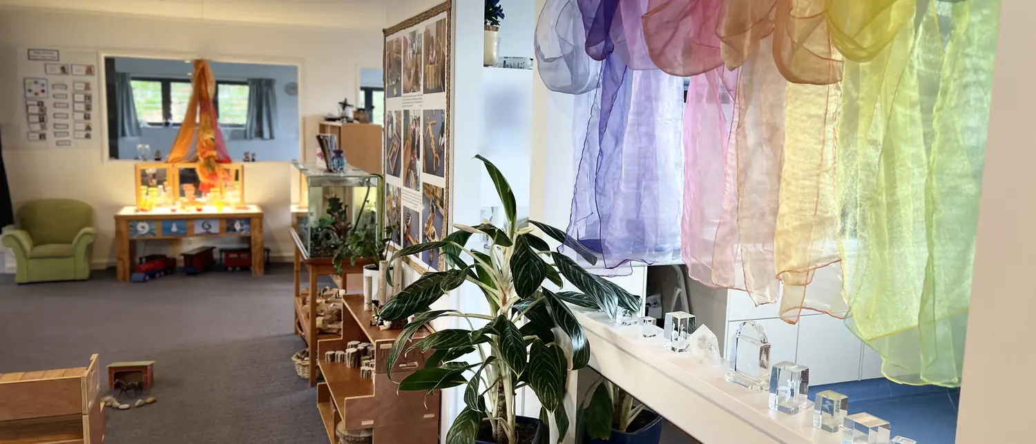 A Magic Garden room with rainbow silks hung in the window, plants, low shelves and a green armchair at the far end.