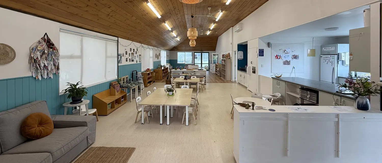A long room at Ponderosa under a timber ceiling and woven pendant lights, with a grey sofa, small tables and chairs, low shelves against teal panelling and a white bench.