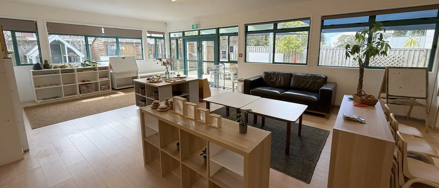 A long room at Anzac Street, Takapuna, with teal-framed windows along one wall, a black leather sofa, an easel, low cube shelves of materials and small tables.