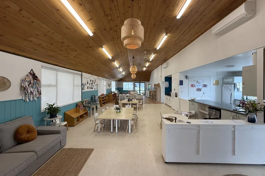 The long room at Ponderosa under a timber ceiling and woven pendant lights, with a grey sofa, small tables and chairs, low shelves against teal panelling and a white bench.