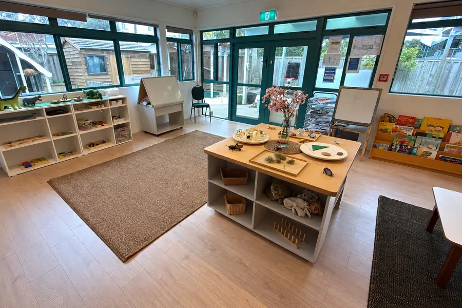 A room at Anzac Street, Takapuna, with teal-framed windows and glass doors to the yard, a discovery table with cherry blossom, low white shelves and a rack of picture books.