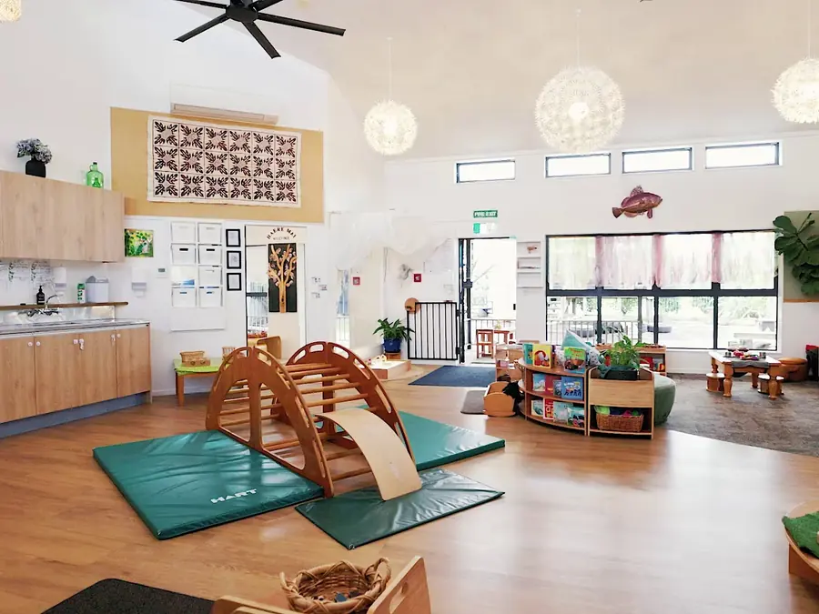 A room with a wooden climbing arch on green mats, low shelves and a kitchen bench along the wall.