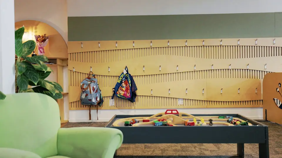 Bags on hooks along a curved wooden wall, with a green armchair and a train table.