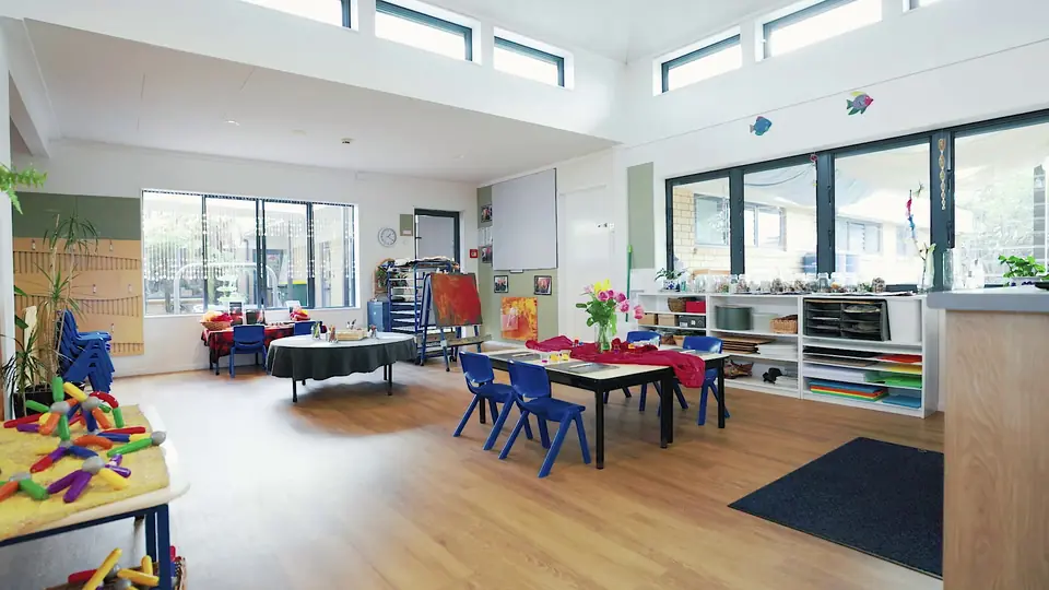 A bright room of small tables and chairs, with vases of flowers and shelves of art materials.