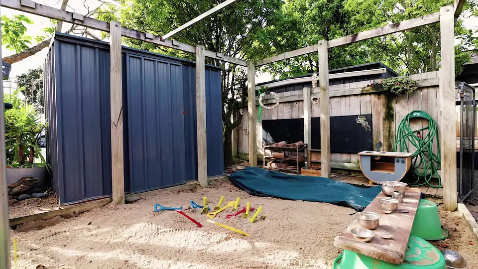 The sandpit and a mud kitchen beside a blue shed, under a timber pergola.
