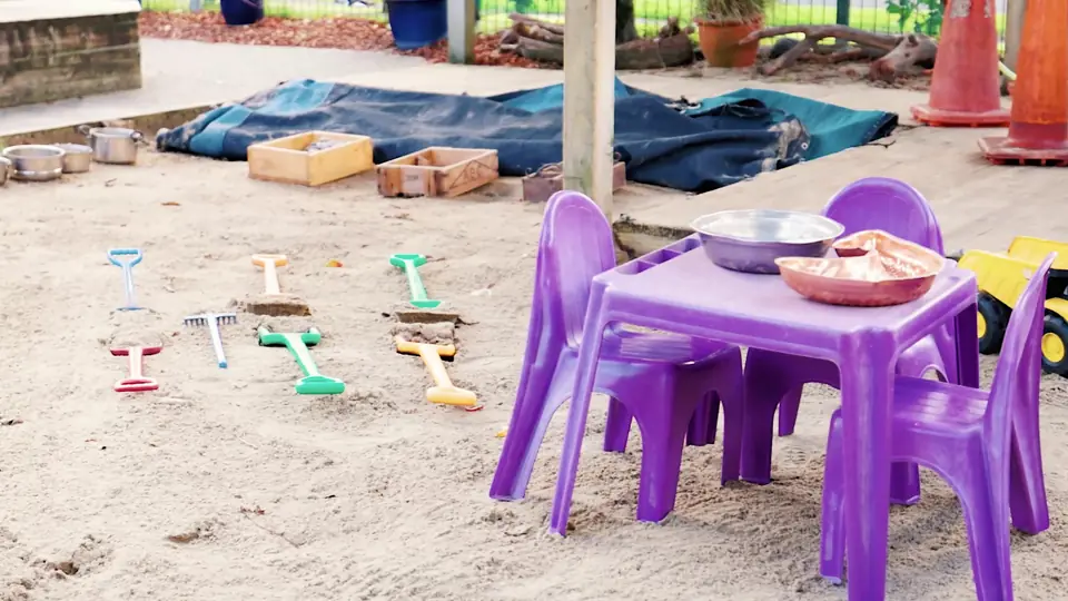 A purple table and chairs set up in the sandpit, with spades and buckets on the sand.
