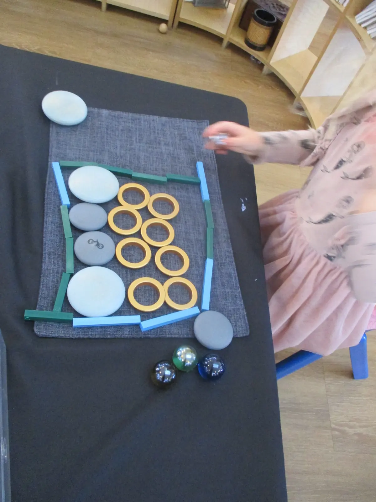Wooden rods, rings and stones laid out on a mat, with a child's arm reaching in.