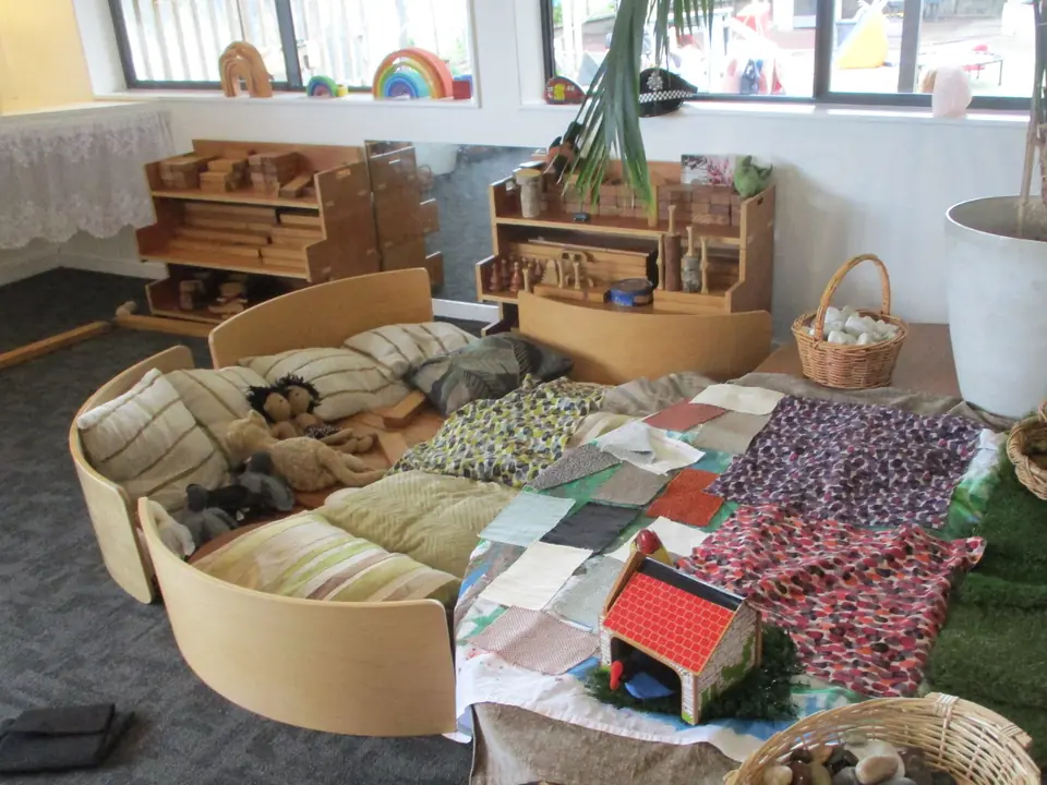 A cushioned nest of curved wood and fabric on the carpet, with patterned cloths and a small wooden house.