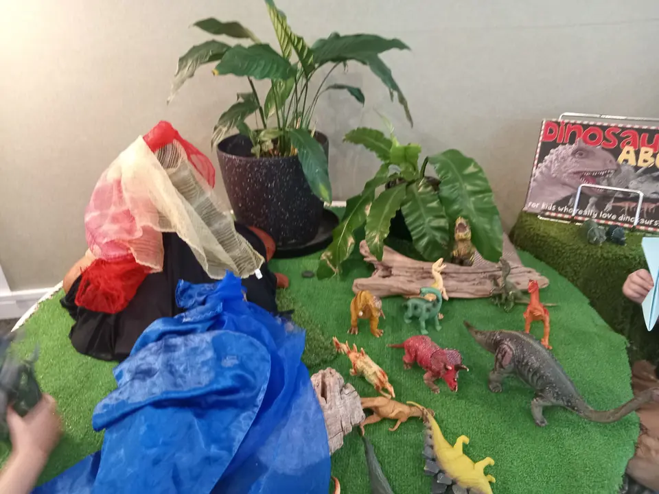 Toy dinosaurs among potted plants on a patch of turf, beside a draped blue cloth.