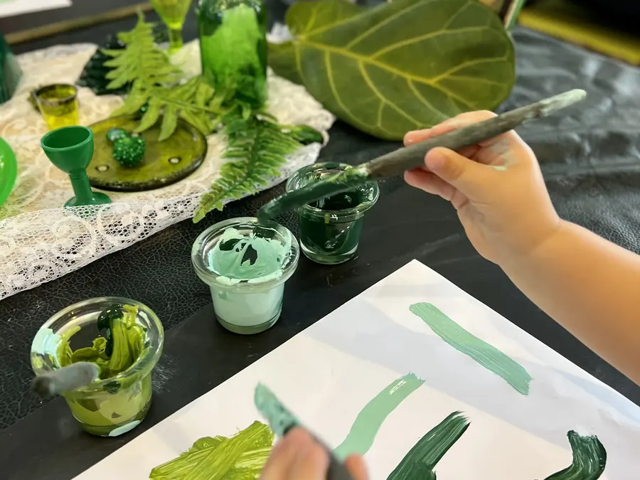 A child's hand painting green strokes on white paper, beside glass pots of paint, ferns and a leaf.
