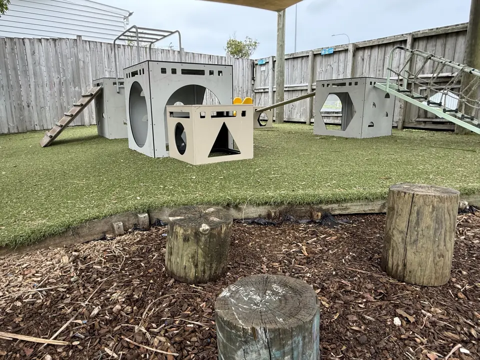 A turf yard with white play cubes cut with round and triangle windows, a wooden ramp, tree stumps in a bark garden and a climbing frame along the fence.