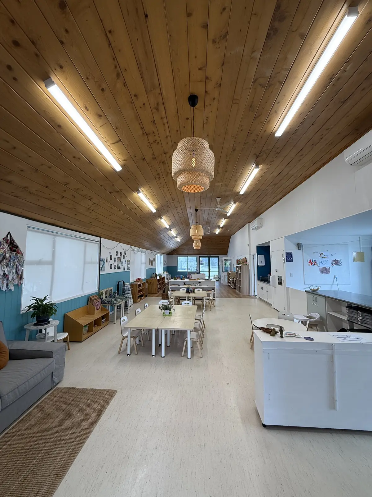A long room under a timber ceiling and woven pendant lights: a grey sofa, small tables and chairs, low shelves against teal panelling and a white bench.