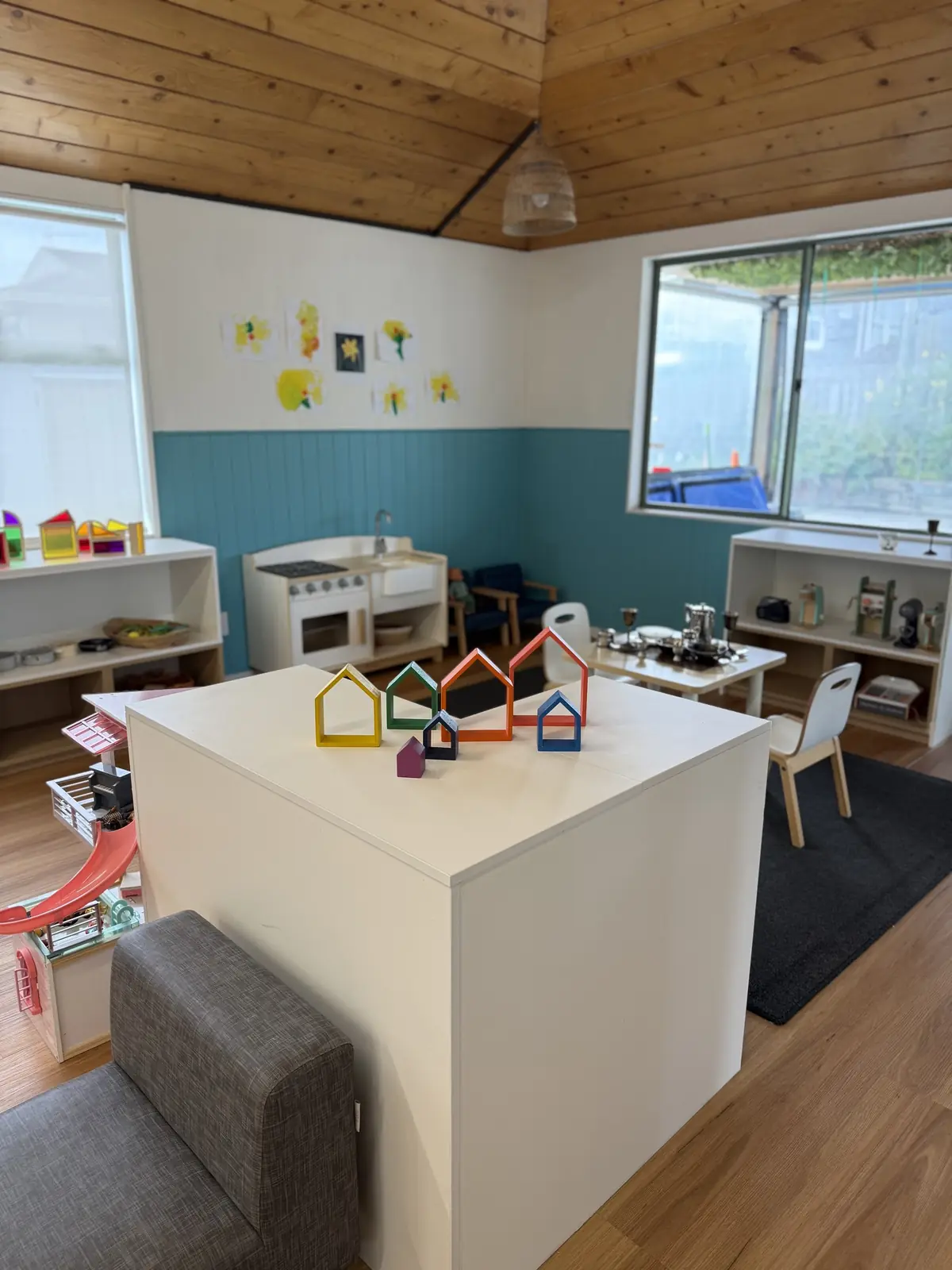 A white table holding rainbow wooden houses, in front of a play kitchen, low shelves and a window.