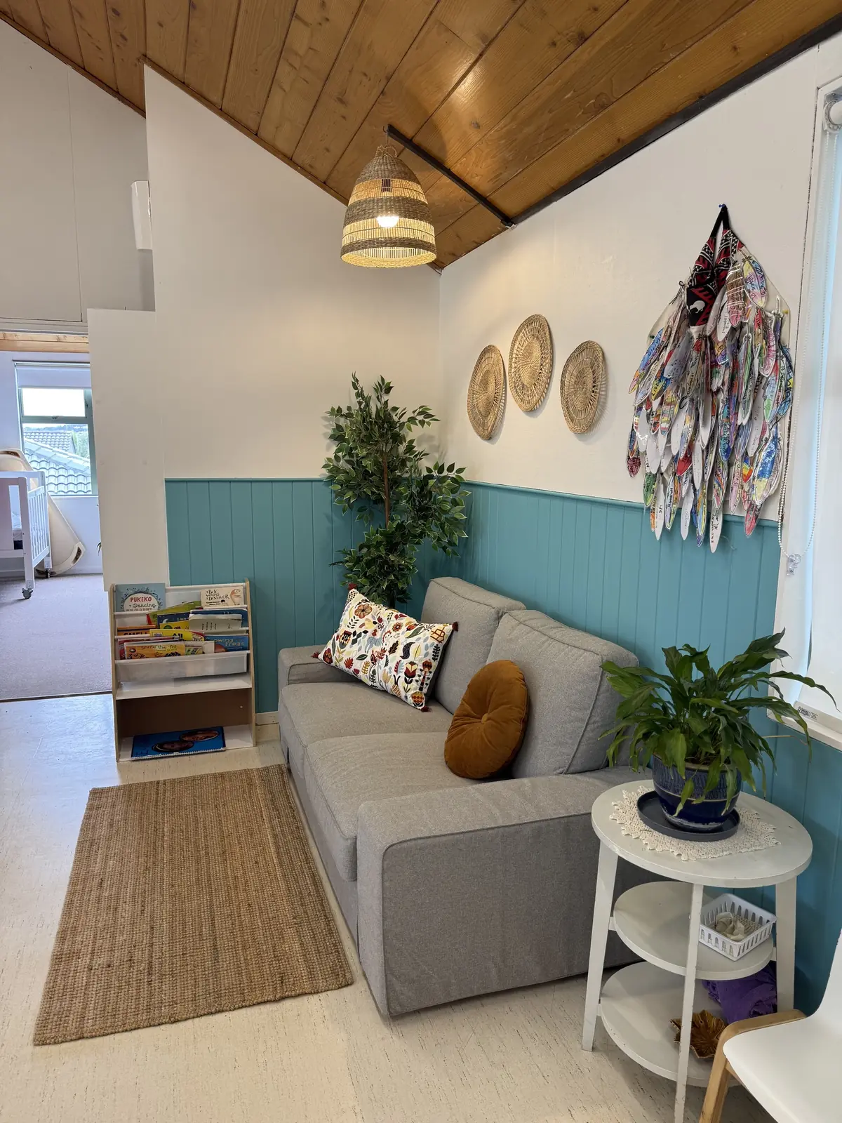 A grey sofa with patterned cushions on a jute rug, a book rack, plants, woven baskets on the wall and a fabric hanging above teal panelling.