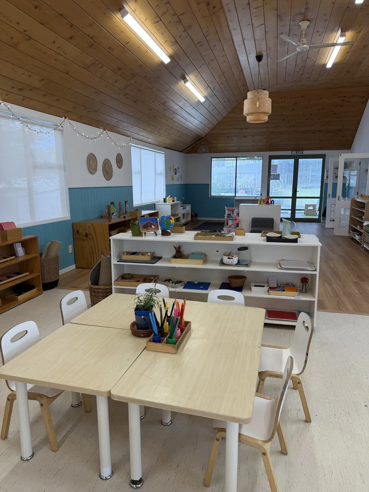Two small tables pushed together with a pot of coloured pencils, in front of a low white shelf of materials and glass doors.