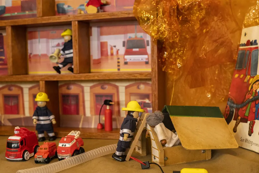 A small-world fire station laid out on a table: wooden firefighter figures in yellow helmets, red toy fire engines, orange cellophane flames and a picture book about a fire engine.