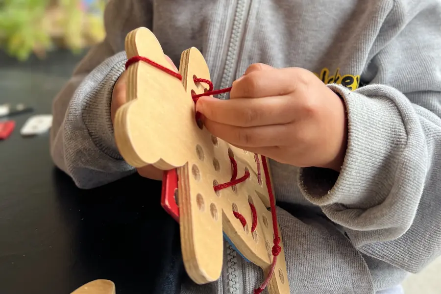 A child in a grey hoodie threads red cord through the holes of a wooden lacing shape.