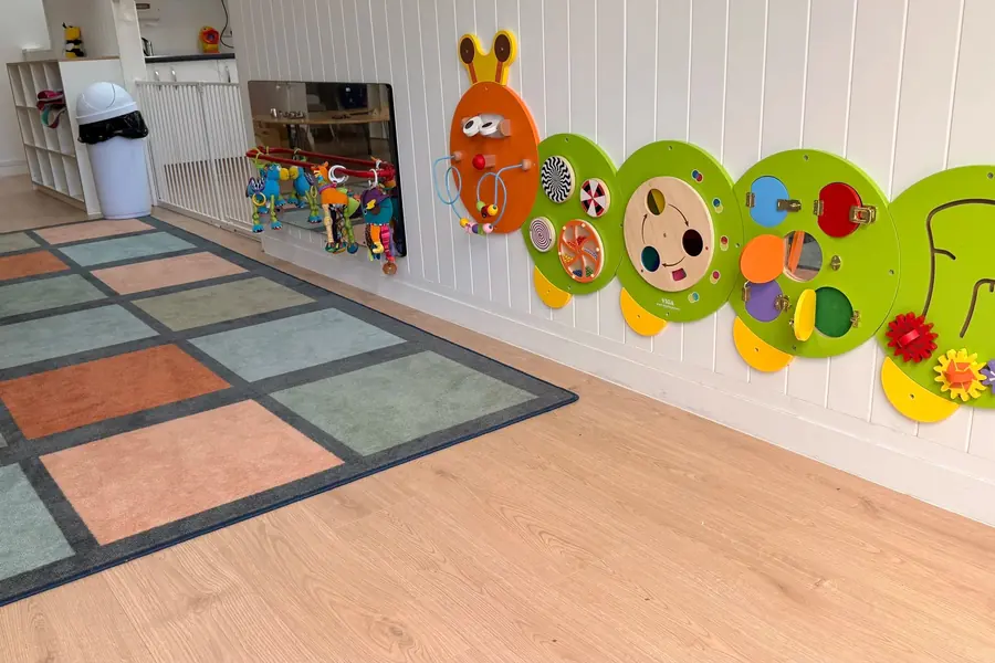The babies’ room at Anzac Street, Takapuna: a green caterpillar of activity boards along the wall, a low mirror with toys hanging in front of it, and a patchwork rug on the wooden floor.