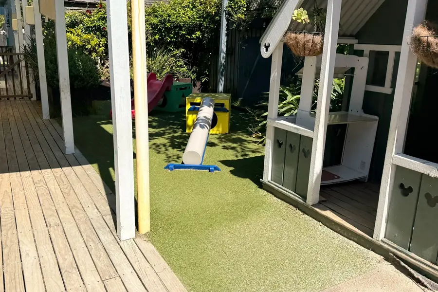 The side yard at Anzac Street, Takapuna: a wooden playhouse with hanging baskets, a balance beam on the turf and a red slide behind, beside a covered deck.