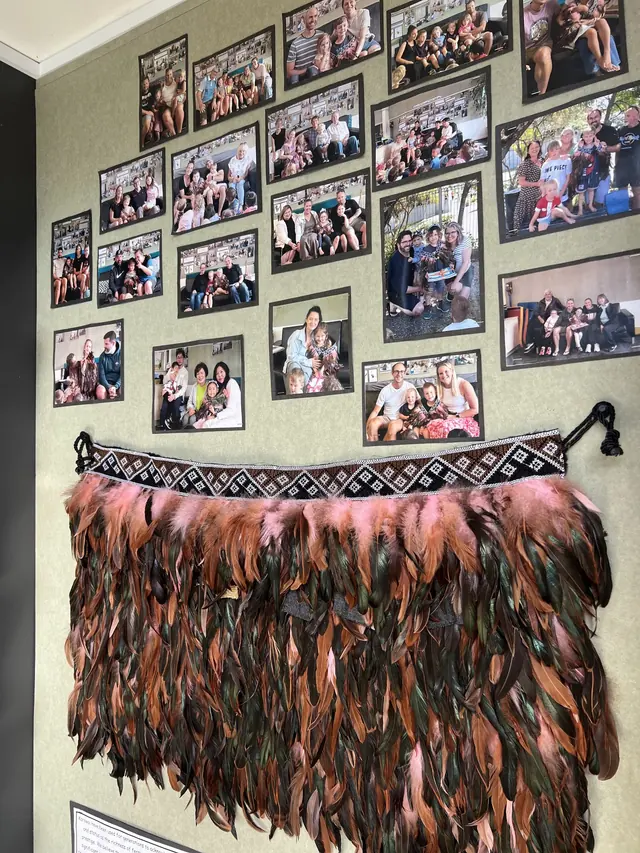 A wall of framed family photographs above a feathered korowai at Northcross.