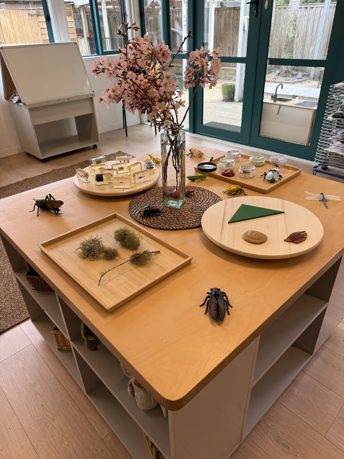 A discovery table with cherry blossom in a vase, toy insects, insects set in clear resin and a tray of dried moss.