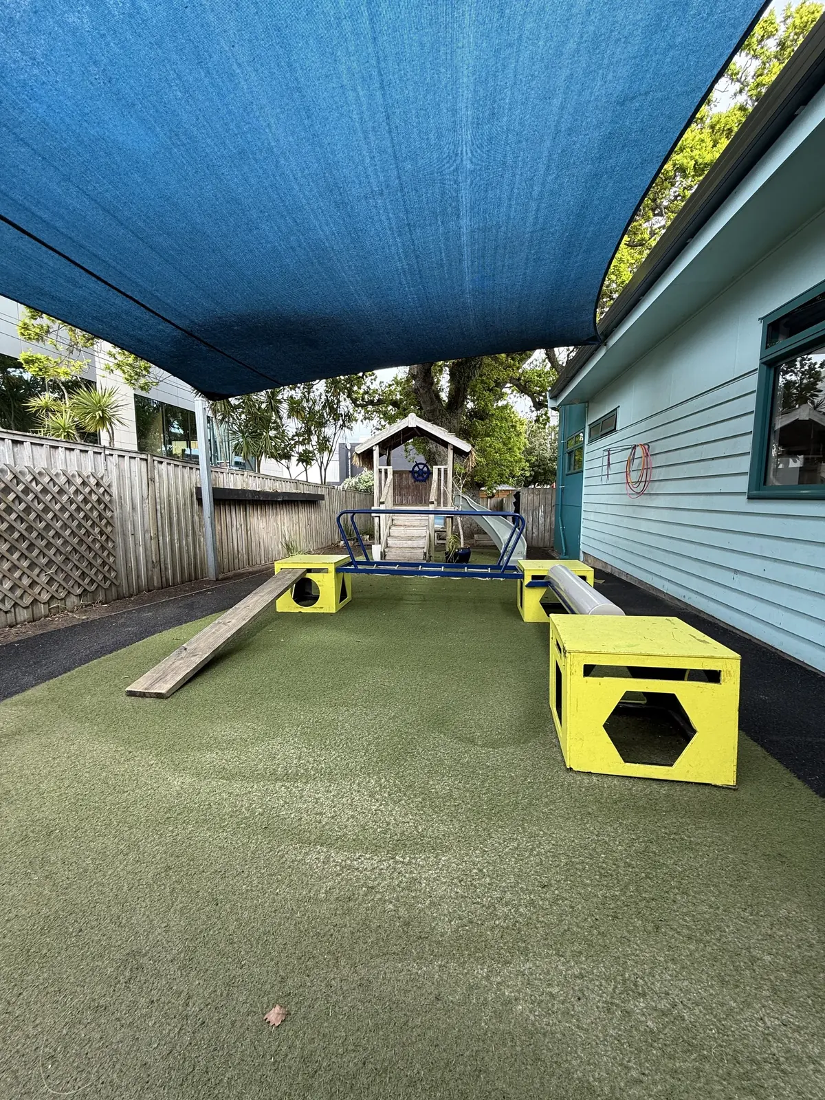 A turf yard under a blue shade sail, with yellow play boxes, a balance plank and a playhouse beyond.