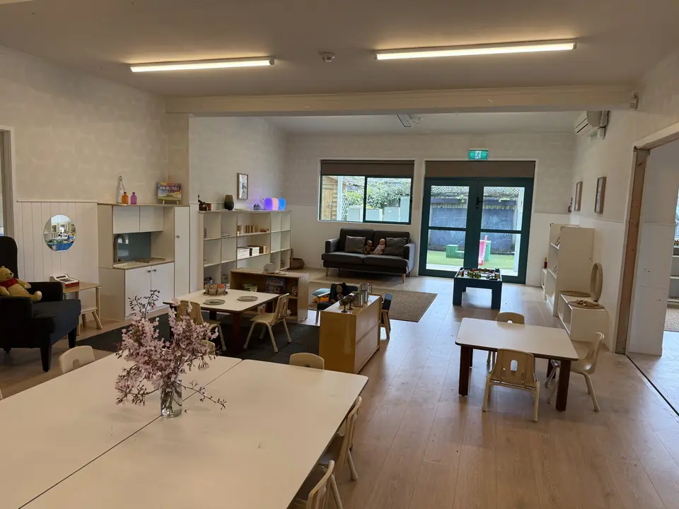 Two rooms opened into one, with a white table and a vase of blossom in front, small tables and chairs, white shelving and glass doors to the yard.