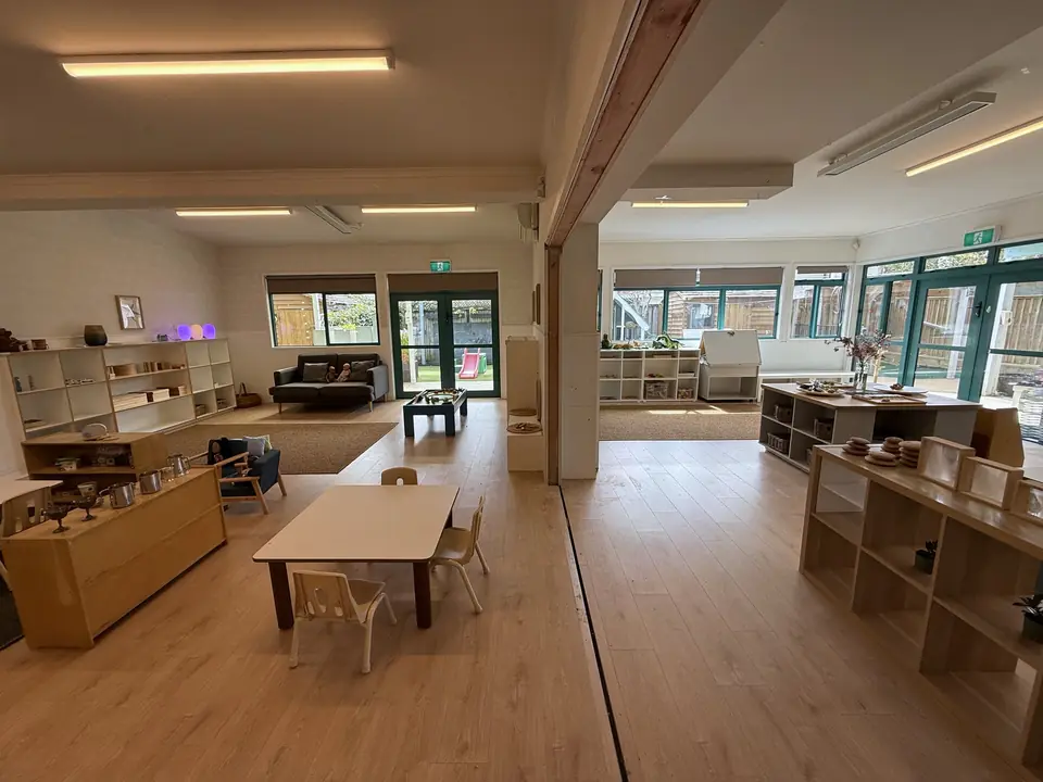 Low wooden shelves of materials and a small table and chairs, looking through a wide opening to a room with teal-framed windows.