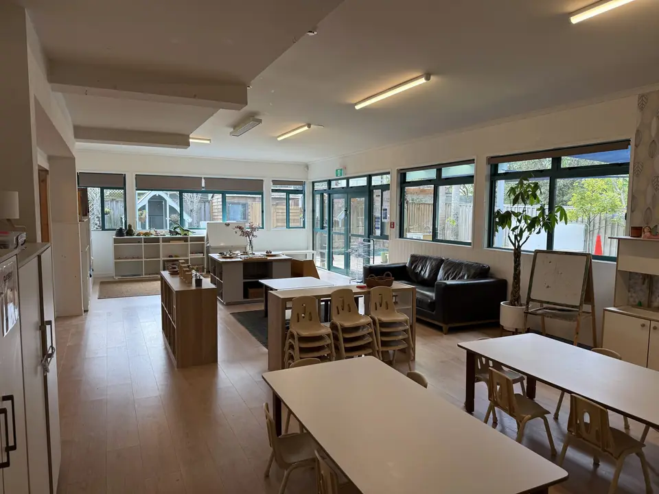 Small tables and stacked chairs in a long room with teal-framed windows, a black leather sofa, a plant and an easel.