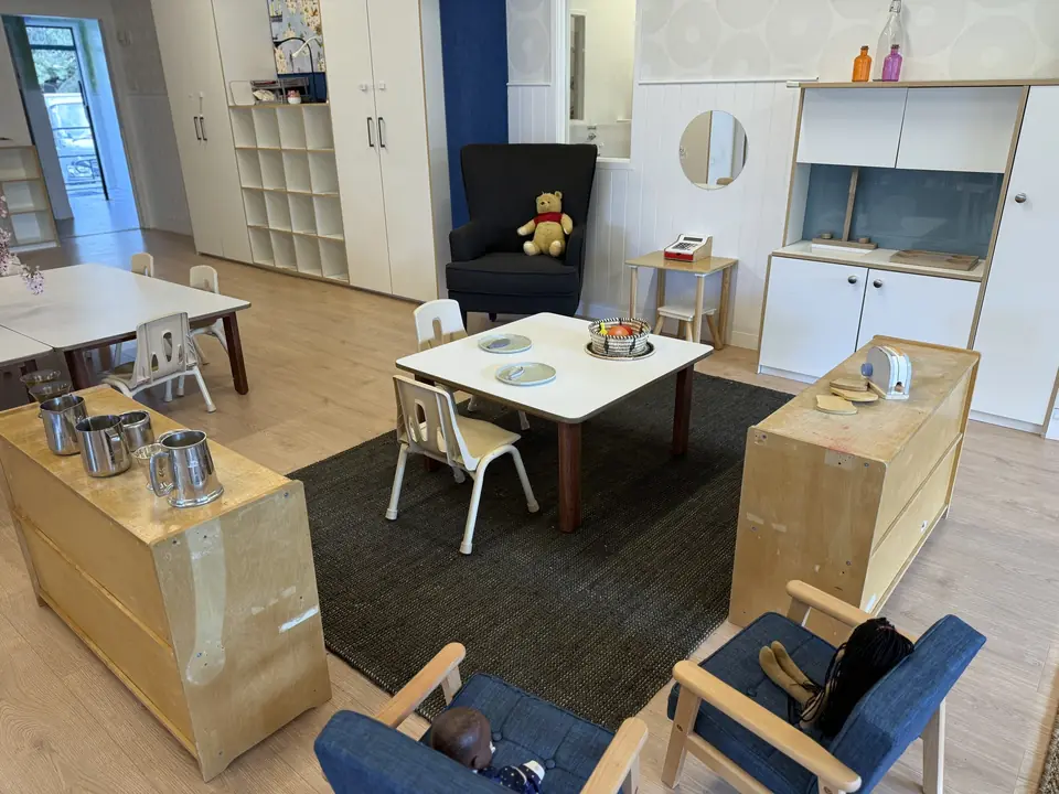A small table set with plates and a basket of fruit, blue armchairs, a black armchair with a teddy bear, and a play kitchen.
