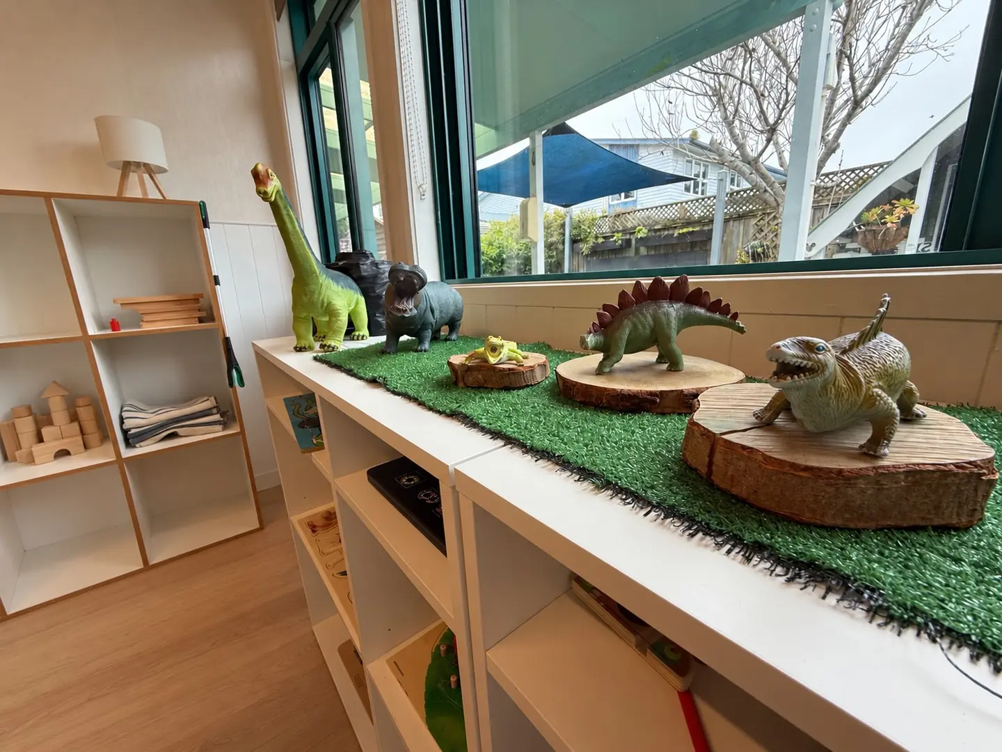 Toy dinosaurs on turf and slices of tree trunk along a low white shelf, under a window looking out to the yard.