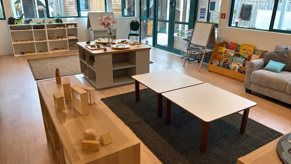 A long room with teal-framed windows and doors, a low table of wooden blocks and frames, two white tables on a dark rug, an easel, a book rack and a sofa.