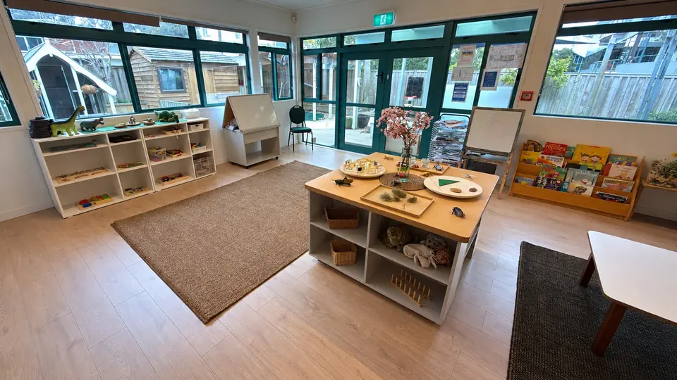 A room with teal-framed windows and glass doors to the yard, a discovery table with cherry blossom, low white shelves, an easel and a rack of picture books.