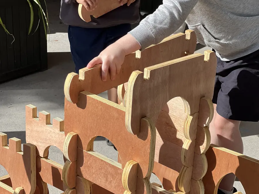 A teacher and a child build together with large wooden castle pieces, their hands on the same wall.