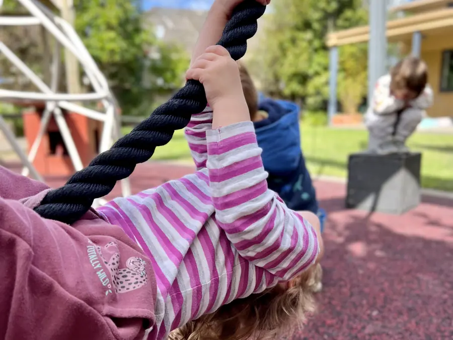 A child in a striped top swings on a thick rope in the playground, seen from behind.